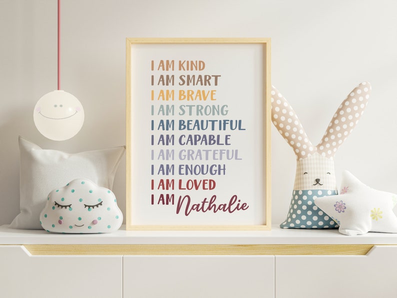 Op de afbeelding: Een ingelijst print met een witte achtergrond en kleurrijke tekst die luidt "Ik ben aardig, ik ben slim, ik ben dapper, ik ben sterk, ik ben mooi, ik ben bekwaam, ik ben dankbaar, ik ben genoeg, ik ben geliefd, ik ben Nathalie". De print staat op een plank met een wit wolkenvormig kussen met groene stippen en een grijs konijnvormig kussen met blauwe stippen.