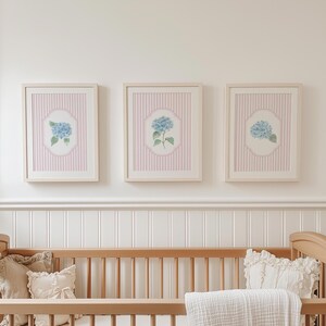 May include: Three framed floral art prints. Each print features a blue hydrangea flower on a white and pink striped background. The frames are light-coloured wood. The prints are displayed above a wooden cot with pillows and a blanket.