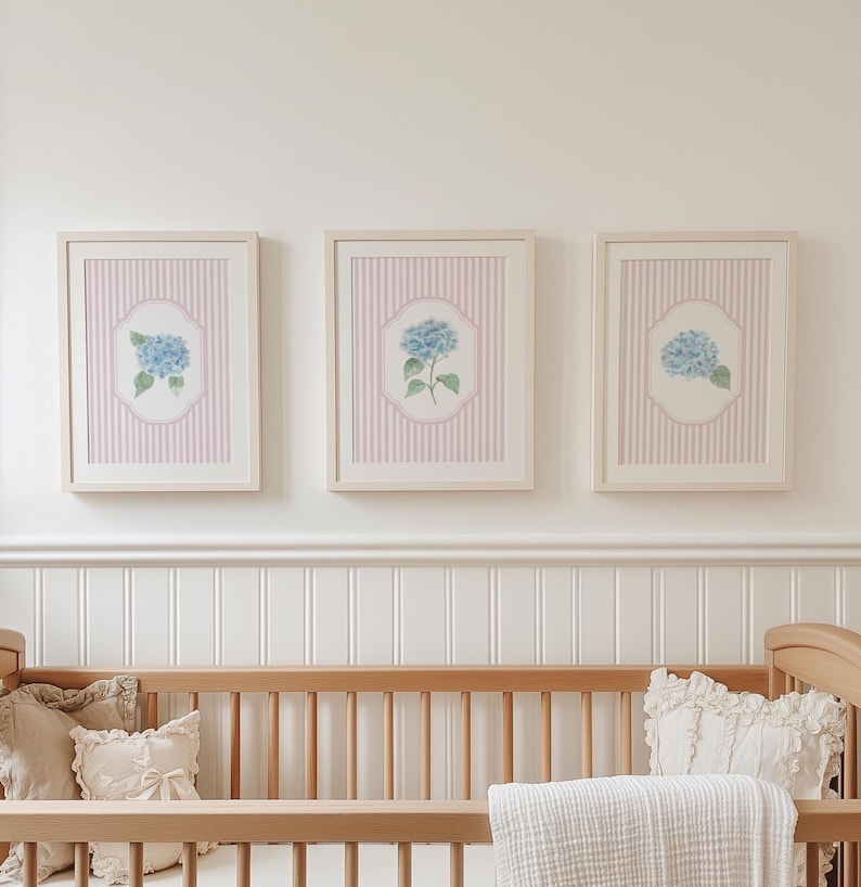 May include: Three framed floral art prints. Each print features a blue hydrangea flower on a white and pink striped background. The frames are light-coloured wood. The prints are displayed above a wooden cot with pillows and a blanket.
