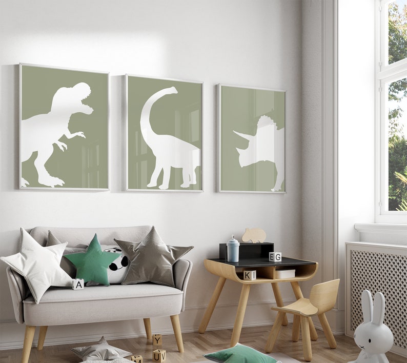May include: Three framed prints of white dinosaur silhouettes on a sage green background. The prints are hanging on a white wall above a light gray couch with green and gray throw pillows. A wooden table with a black top and a wooden chair are in the foreground.