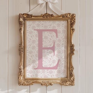 May include: A framed print featuring a large pink letter "E" against a floral patterned background. The ornate gold frame is suspended by a white ribbon tied in a bow. The print is hung against a white wooden wall.