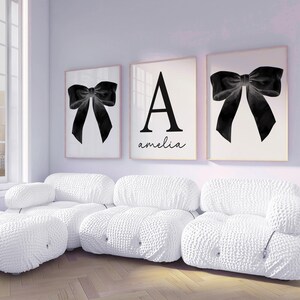 Peut inclure: Trois impressions encadr&eacute;es avec un motif de n&oelig;ud noir et une grande lettre "A" avec le nom "amelia" en &eacute;criture cursive. Les impressions sont au-dessus d'un canap&eacute; sectionnel blanc textur&eacute; dans une pi&egrave;ce lumineuse.