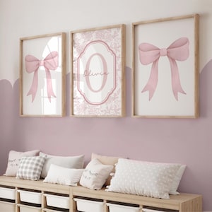Puede incluir: Tres impresiones art&iacute;sticas enmarcadas con un acabado de madera clara. Dos impresiones presentan una ilustraci&oacute;n de lazo rosa, mientras que la impresi&oacute;n central muestra el nombre "Olivia" en cursiva dentro de un marco floral rosa. Las impresiones se exhiben sobre un banco de madera.