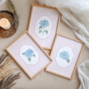 May include: Three framed art prints with blue hydrangea illustrations on a pink and white striped background. The frames are light wood. The prints are displayed on a neutral surface, with candles and decorative elements.