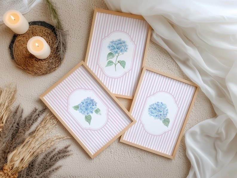 May include: Three framed art prints with blue hydrangea illustrations on a pink and white striped background. The frames are light wood. The prints are displayed on a neutral surface, with candles and decorative elements.