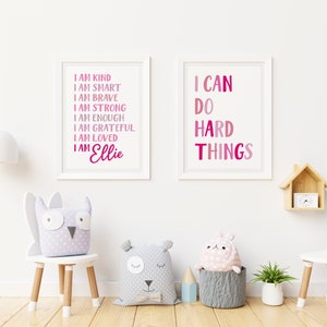 Pu&ograve; includere: Due stampe incorniciate con testo rosa e bianco su sfondo bianco. La stampa a sinistra recita "Sono gentile, sono intelligente, sono coraggioso, sono forte, sono abbastanza, sono grato, sono amato, sono Ellie". La stampa a destra recita "Posso fare cose difficili".