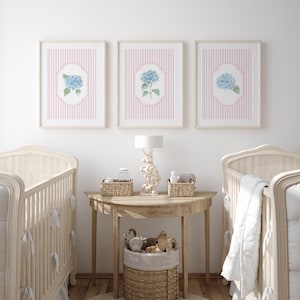 May include: Three framed prints with blue hydrangea flowers on a pink striped background hang above a wooden table with a lamp and two cots. A woven basket holds toys. The room has a soft, neutral colour palette.