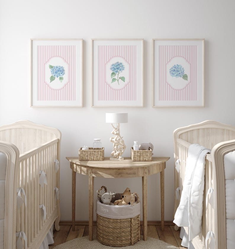May include: Three framed prints with blue hydrangea flowers on a pink striped background hang above a wooden table with a lamp and two cots. A woven basket holds toys. The room has a soft, neutral colour palette.