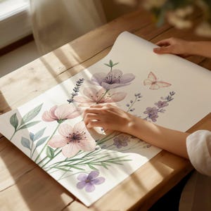 May include: A floral print on a rolled-up paper. The design features watercolor illustrations of pink and purple flowers, green leaves, and a butterfly. The paper is being held on a wooden surface.