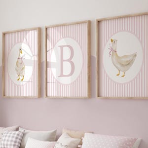 May include: Three framed prints with a nursery theme. Each print features a watercolor illustration of a goose with a pink ribbon, set against a pink and white striped background. One print has the letter "B". The frames are light wood.