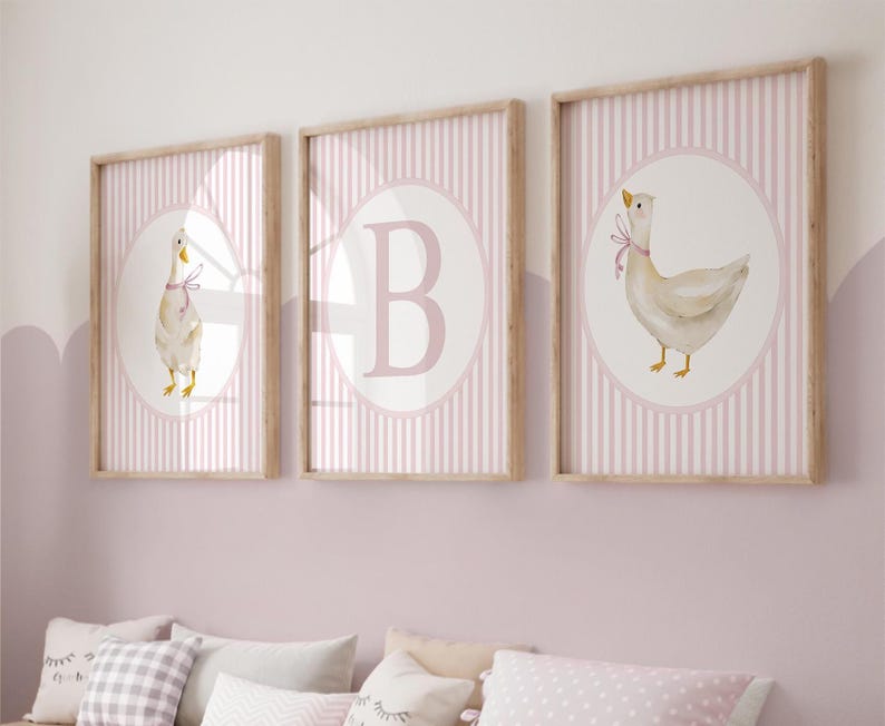 May include: Three framed prints with a nursery theme. Each print features a watercolor illustration of a goose with a pink ribbon, set against a pink and white striped background. One print has the letter "B". The frames are light wood.