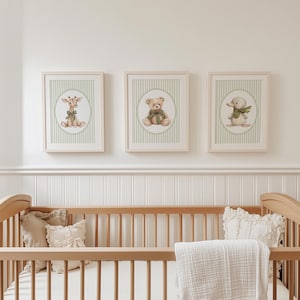 May include: Three framed nursery art prints. Each print features a different animal: a giraffe, a teddy bear, and a duck. The prints have a light green striped background and a light wood frame. The art is displayed above a wooden cot.