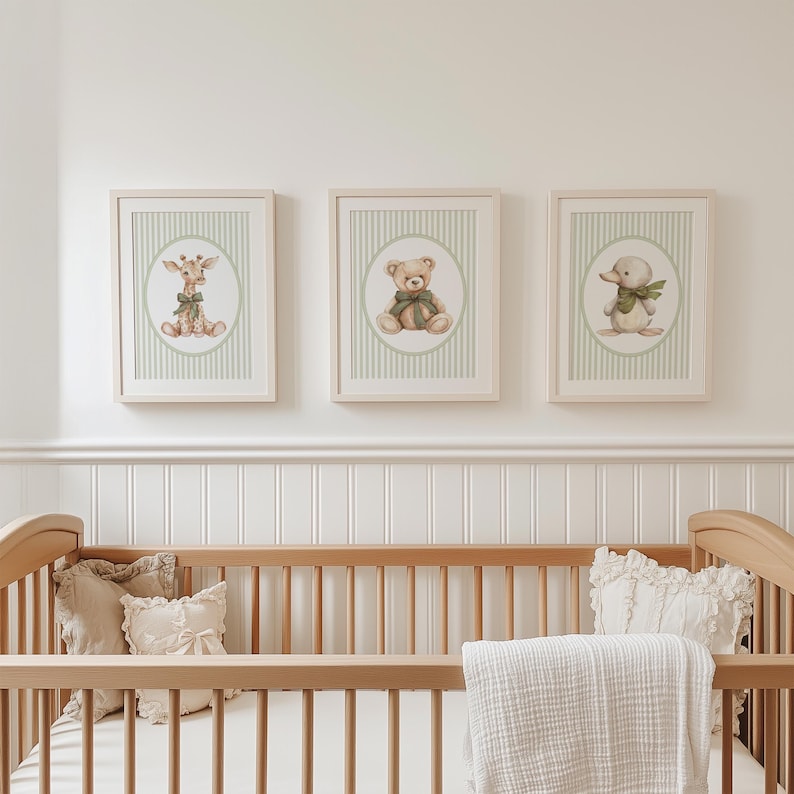May include: Three framed nursery art prints. Each print features a different animal: a giraffe, a teddy bear, and a duck. The prints have a light green striped background and a light wood frame. The art is displayed above a wooden cot.