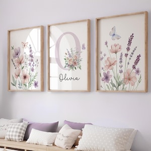 May include: Three framed floral art prints. Each print features watercolor flowers in shades of pink, purple, and lavender, with butterflies. The center print has the letter "O" and the name "Olivia". The frames are light brown.