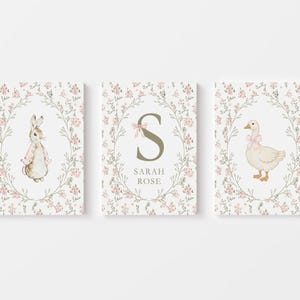 May include: Three framed art prints featuring a bunny, a goose, and the letter "S" with the name "SARAH ROSE". Each print has a floral wreath border with pink flowers and green leaves. The bunny and goose wear pink bows.