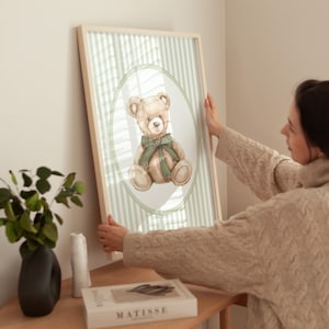 May include: A framed print with a watercolour illustration of a teddy bear wearing a green bow. The bear is set against a light green striped background within an oval frame. The frame is light wood-toned.