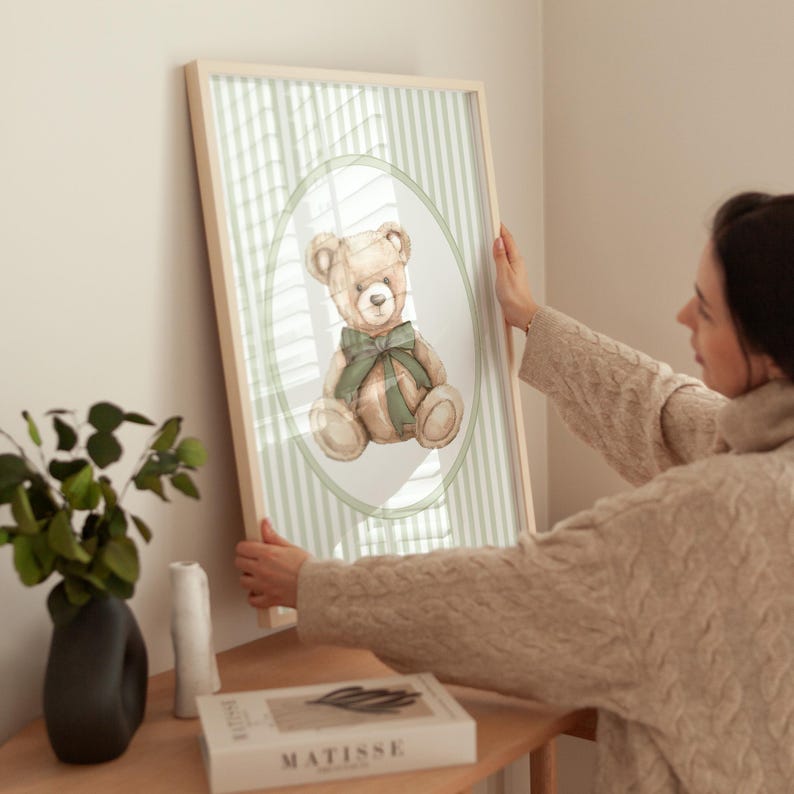 May include: A framed print with a watercolour illustration of a teddy bear wearing a green bow. The bear is set against a light green striped background within an oval frame. The frame is light wood-toned.
