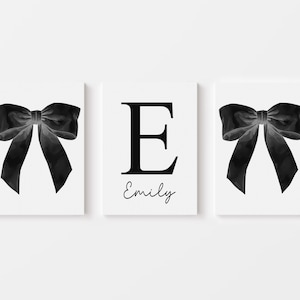 Peut inclure: Triptyque d'impressions d'art blanches. Deux impressions pr&eacute;sentent un n&oelig;ud noir &agrave; l'aquarelle. L'impression centrale affiche la lettre "E" en noir, avec le nom "Emily" en cursive en dessous. Le fond est blanc.