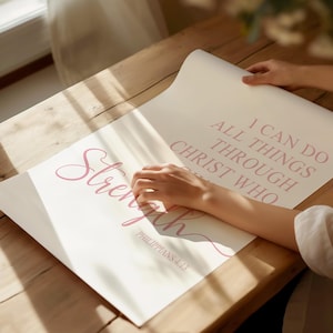May include: A light-colored poster with the word "Strength" in a cursive font and the text "I can do all things through Christ who" in a serif font. The text "Philippians 4:13" is also visible. The poster is being held on a wooden surface.