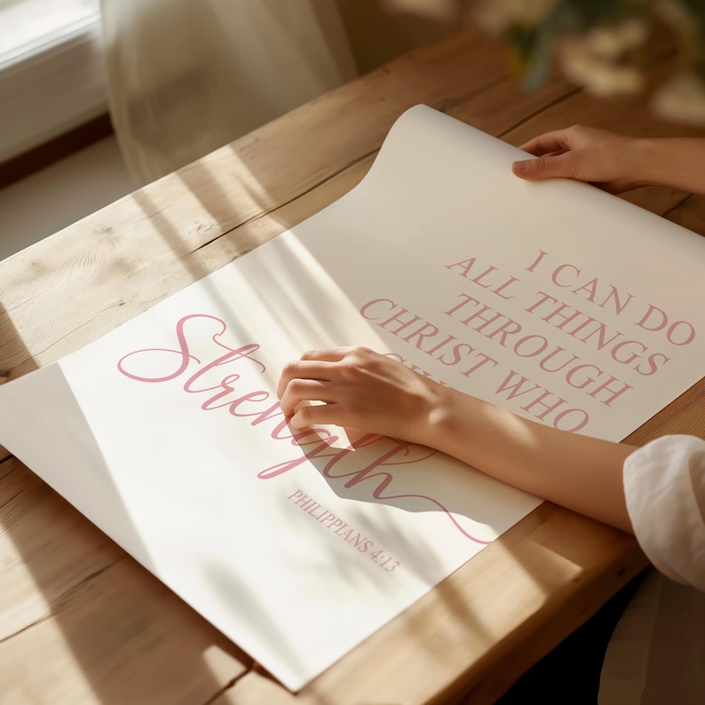 May include: A light-colored poster with the word "Strength" in a cursive font and the text "I can do all things through Christ who" in a serif font. The text "Philippians 4:13" is also visible. The poster is being held on a wooden surface.