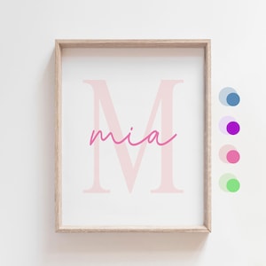 May include: A framed print with a pink letter M and the word "mia" written in pink cursive. The frame is light brown wood.