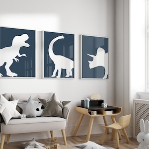 May include: Three framed prints of white dinosaur silhouettes on a dark blue background. The prints are hanging on a white wall above a light gray couch with pillows and a small wooden table and chair.