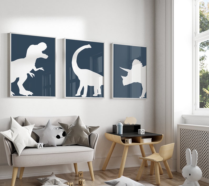 May include: Three framed prints of white dinosaur silhouettes on a dark blue background. The prints are hanging on a white wall above a light gray couch with pillows and a small wooden table and chair.