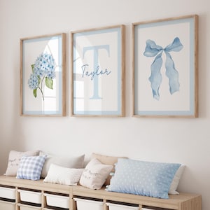 May include: Three framed prints with light blue accents. One features a blue hydrangea, another a blue letter "T" with the name "Taylor", and the third a blue watercolor bow. The frames are light wood.