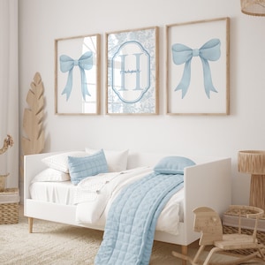 Puede incluir: Tres impresiones enmarcadas con ilustraciones de lazo azul claro y un monograma. La impresi&oacute;n central presenta el nombre "Harper". La obra de arte est&aacute; sobre una cama blanca con ropa de cama y almohadas azul claro. Un caballito de madera y una l&aacute;mpara tambi&eacute;n est&aacute;n en la habitaci&oacute;n.