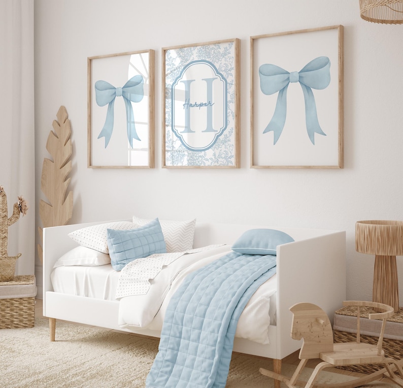 Puede incluir: Tres impresiones enmarcadas con ilustraciones de lazo azul claro y un monograma. La impresi&oacute;n central presenta el nombre "Harper". La obra de arte est&aacute; sobre una cama blanca con ropa de cama y almohadas azul claro. Un caballito de madera y una l&aacute;mpara tambi&eacute;n est&aacute;n en la habitaci&oacute;n.