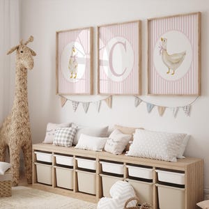 May include: Three framed prints with a pink striped background and a white circle featuring a goose illustration. A large woven giraffe and a storage bench with pillows and bins complete the nursery decor.