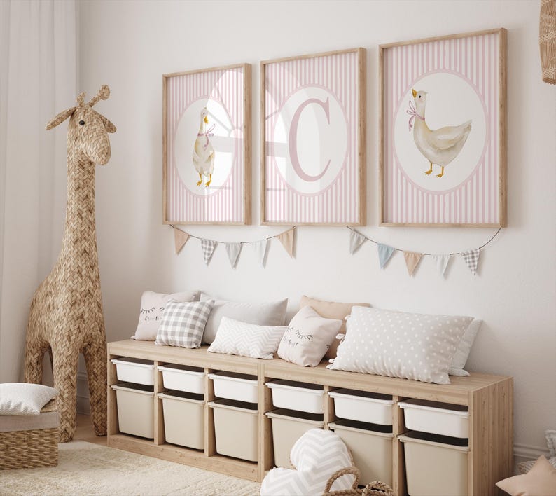 May include: Three framed prints with a pink striped background and a white circle featuring a goose illustration. A large woven giraffe and a storage bench with pillows and bins complete the nursery decor.