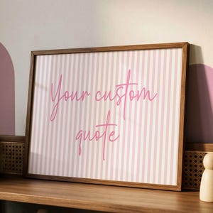 May include: A rectangular wooden frame containing a print with vertical light pink and white stripes. The text "Your custom quote" is written in a cursive, dark pink font across the center of the striped background within the frame.