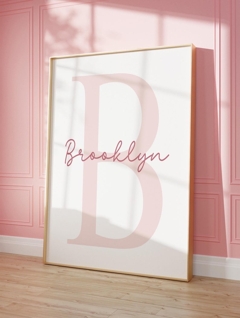 May include: A framed print featuring a large, light pink letter "B" with the name "Brooklyn" in a cursive font below it. The print has a white background and a gold frame. The artwork is displayed against a pink wall.