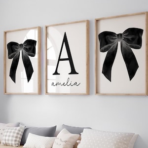 Peut inclure: Trois cadres muraux. Deux pr&eacute;sentent une illustration d'un n&oelig;ud noir sur fond blanc. Le cadre central affiche la lettre noire "A" et le pr&eacute;nom "amelia" en &eacute;criture cursive. Les cadres sont en bois clair.