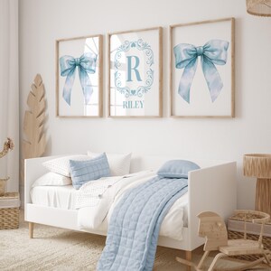 May include: Three framed prints with a blue watercolor bow, a monogram with the letter R and the name Riley, and another blue watercolor bow. The prints are hanging on a white wall above a white daybed with blue bedding.