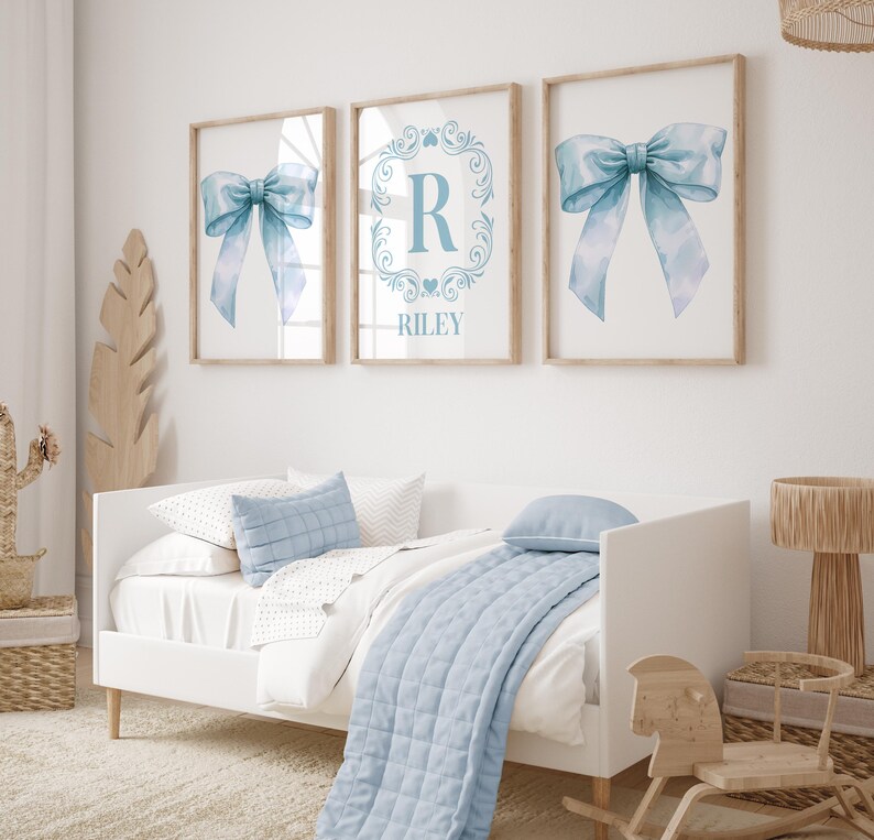 May include: Three framed prints with a blue watercolor bow, a monogram with the letter R and the name Riley, and another blue watercolor bow. The prints are hanging on a white wall above a white daybed with blue bedding.