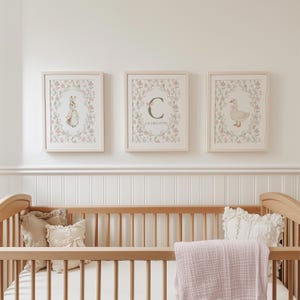 May include: Three framed nursery art prints featuring a bunny, a goose, and a floral initial. The prints are in light wood frames and are decorated with pink and green floral wreaths. The prints are displayed above a wooden crib with pillows and a pink blanket.