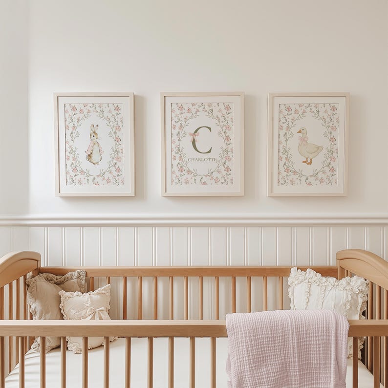 May include: Three framed nursery art prints featuring a bunny, a goose, and a floral initial. The prints are in light wood frames and are decorated with pink and green floral wreaths. The prints are displayed above a wooden crib with pillows and a pink blanket.