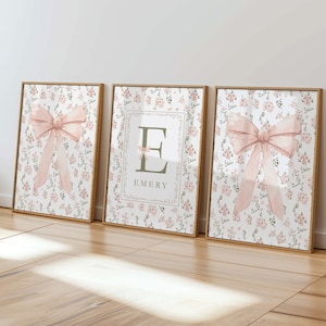 May include: Three framed art prints with a floral pattern and a light pink bow design. The center print features the name "Emery" and the letter "E". The frames are a light brown color.