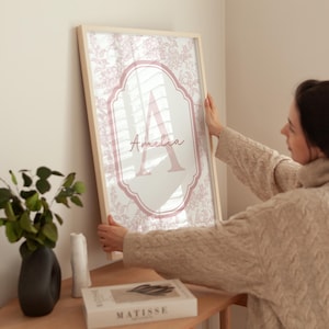 Puede incluir: Un estampado enmarcado con un dise&ntilde;o floral en rosa y blanco. El estampado tiene un borde rosa con la letra "A" y el nombre "Amelia" en escritura cursiva. El marco es de madera clara.