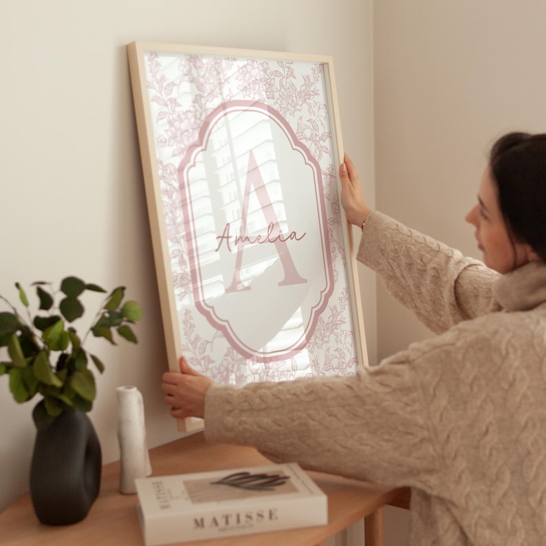 Puede incluir: Un estampado enmarcado con un dise&ntilde;o floral en rosa y blanco. El estampado tiene un borde rosa con la letra "A" y el nombre "Amelia" en escritura cursiva. El marco es de madera clara.
