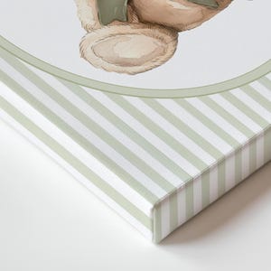 May include: A canvas print featuring a watercolour illustration of a teddy bear with a green bow. The canvas edges are wrapped with a light green and white striped pattern, creating a soft, vintage aesthetic.