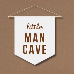 Peut inclure: Banni&egrave;re blanche avec du texte brun qui dit "little MAN CAVE" accroch&eacute;e &agrave; un mur brun.