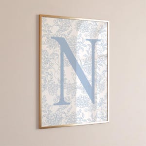 May include: Framed wall art featuring a large, light blue letter "N" overlaid on a floral pattern. The background has a cream color with light blue floral designs. The frame is gold.