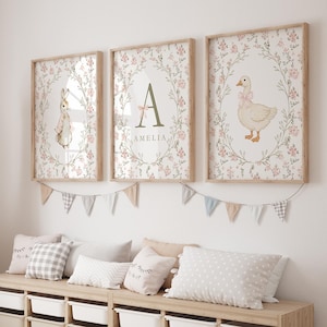 May include: Three framed nursery art prints. One features a bunny, another a letter "A" with the name "Amelia", and the third a goose. All are surrounded by floral wreaths and set against a floral background. A garland of small flags hangs below.