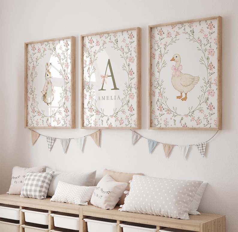 May include: Three framed nursery art prints. One features a bunny, another a letter "A" with the name "Amelia", and the third a goose. All are surrounded by floral wreaths and set against a floral background. A garland of small flags hangs below.