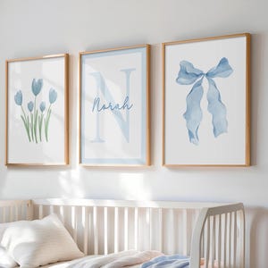 May include: Three framed art prints with watercolor designs. The prints feature a blue bow, blue tulips, and the letter "N" with the name "Norah" in script. The frames are a light wood color, and the art is displayed above a white crib.