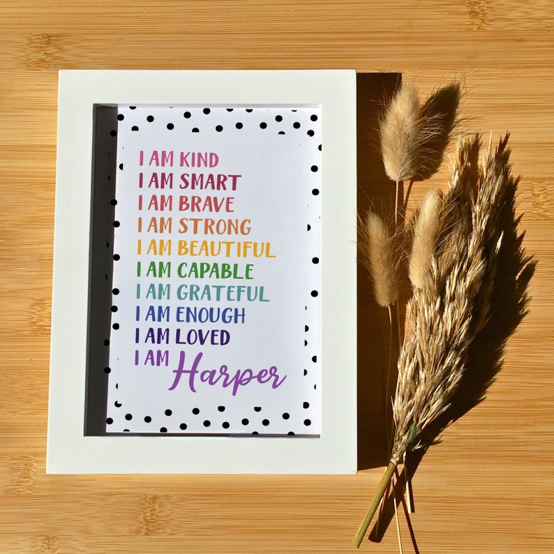 Op de afbeelding: Een wit ingelijst print met een zwart-witte stippenachtergrond. De print heeft een regenboog van kleuren met de tekst "Ik ben aardig, ik ben slim, ik ben dapper, ik ben sterk, ik ben mooi, ik ben bekwaam, ik ben dankbaar, ik ben genoeg, ik ben geliefd, ik ben Harper".