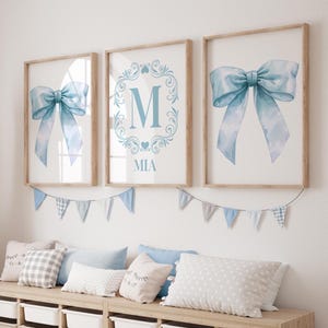 May include: Three framed prints with a blue watercolor bow and the letter M in a decorative frame with the name Mia below. The prints are hanging on a white wall above a wooden bench with pillows.